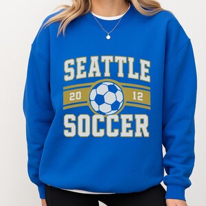 Pode incluir: Um moletom azul royal com as palavras "SEATTLE SOCCER" em letras brancas e douradas. Um gr&aacute;fico de bola de futebol est&aacute; centrado acima da palavra "SOCCER". Os n&uacute;meros "20" e "12" est&atilde;o em ambos os lados da bola.