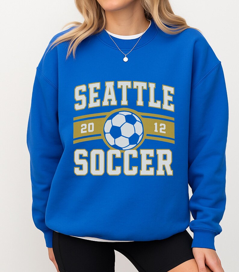 Pode incluir: Um moletom azul royal com as palavras "SEATTLE SOCCER" em letras brancas e douradas. Um gr&aacute;fico de bola de futebol est&aacute; centrado acima da palavra "SOCCER". Os n&uacute;meros "20" e "12" est&atilde;o em ambos os lados da bola.