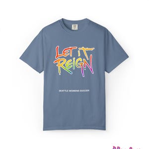 May include: A blue t-shirt with the words "LET IT REIGN" in a rainbow gradient, with "SEATTLE WOMENS SOCCER" below. The shirt is a solid color and has short sleeves.