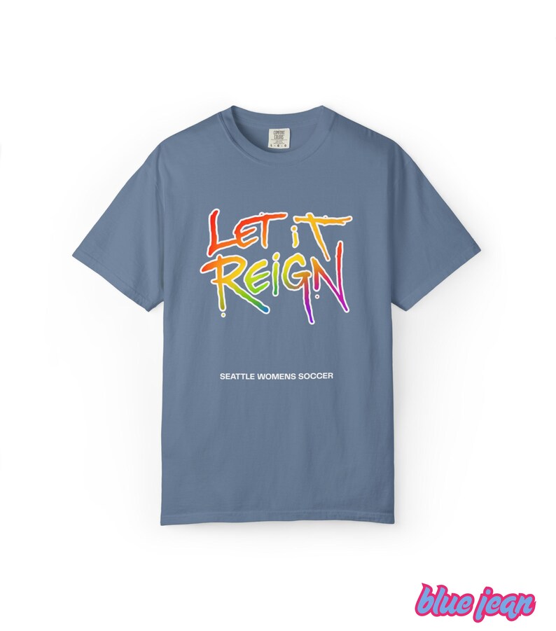 May include: A blue t-shirt with the words "LET IT REIGN" in a rainbow gradient, with "SEATTLE WOMENS SOCCER" below. The shirt is a solid color and has short sleeves.