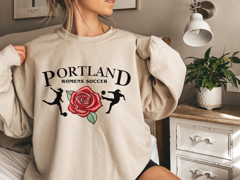 May include: A beige sweatshirt with the text "PORTLAND WOMEN'S SOCCER" and a red rose with two soccer players in black silhouettes.