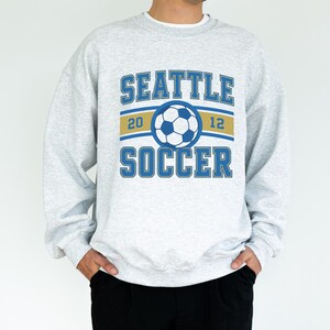 Pode incluir: Camisola cinza claro com as palavras "SEATTLE SOCCER" em letras azuis. Um gr&aacute;fico de bola de futebol est&aacute; centrado acima da palavra "SOCCER". O ano "2012" &eacute; impresso em ambos os lados da bola.
