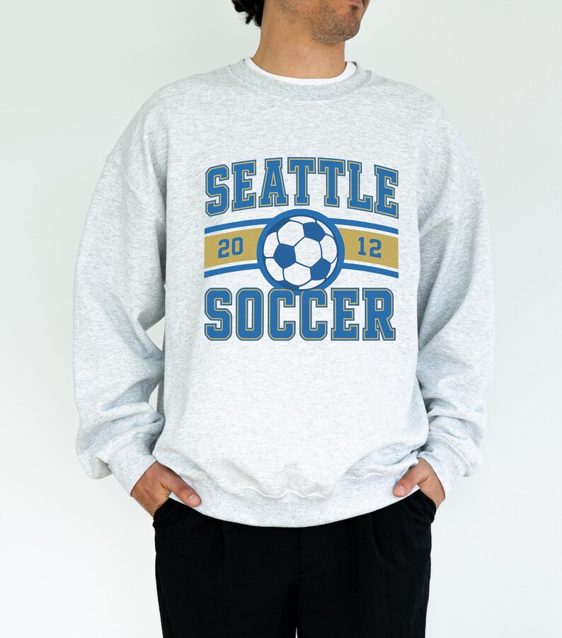 Pode incluir: Camisola cinza claro com as palavras "SEATTLE SOCCER" em letras azuis. Um gr&aacute;fico de bola de futebol est&aacute; centrado acima da palavra "SOCCER". O ano "2012" &eacute; impresso em ambos os lados da bola.