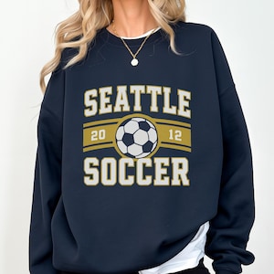 Pode incluir: Camisola azul marinho com as palavras "SEATTLE SOCCER" em letras brancas e douradas. Um gr&aacute;fico de bola de futebol est&aacute; centrado acima da palavra "SOCCER". Os n&uacute;meros "20" e "12" ladeiam a bola.