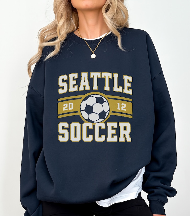 Pode incluir: Camisola azul marinho com as palavras "SEATTLE SOCCER" em letras brancas e douradas. Um gr&aacute;fico de bola de futebol est&aacute; centrado acima da palavra "SOCCER". Os n&uacute;meros "20" e "12" ladeiam a bola.