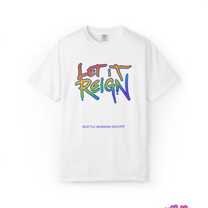 May include: White t-shirt with the text "LET IT REIGN" in rainbow colors. Below the text is "SEATTLE WOMENS SOCCER". The shirt has short sleeves and a crew neck.