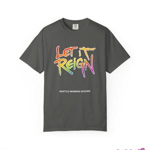 May include: A dark gray t-shirt with the words "Let It Reign" in a rainbow gradient font. Below the text is "Seattle Womens Soccer" in white. The shirt is a crew neck style.