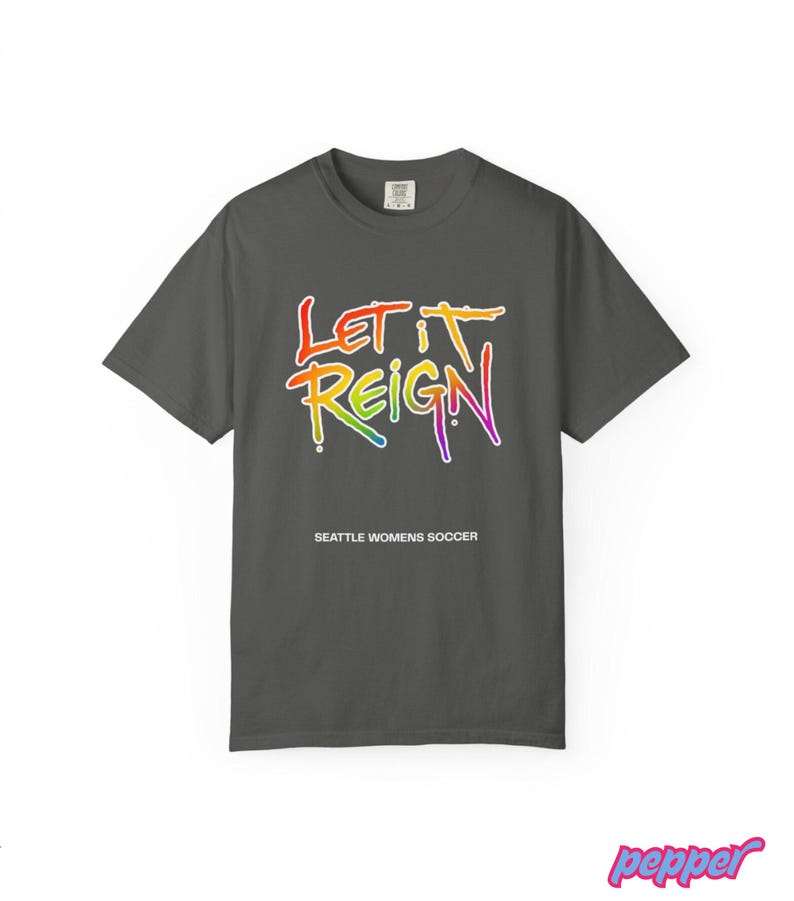 May include: A dark gray t-shirt with the words "Let It Reign" in a rainbow gradient font. Below the text is "Seattle Womens Soccer" in white. The shirt is a crew neck style.