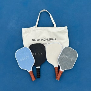 May include: Four pickleball paddles with the word "NALOY" printed on each. The paddles are in a variety of colors and patterns, including blue, black, white, and gray. The paddles are sitting on a blue surface, and a white tote bag with the words "NALOY PICKLEBALL" printed on it is behind the paddles.