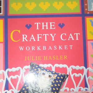 May include: A book cover titled "The Crafty Cat Workbasket" with a pink background and a white bench holding three cat-themed pillows. The cover features pixelated floral and heart designs, and the author's name is Julie Hasler.