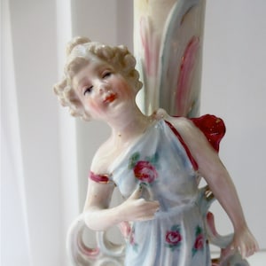May include: A porcelain figurine of a woman in a light blue dress adorned with pink roses. The figurine has a red bow and is part of a lamp base. The base features ornate scrollwork and floral accents, with a cream, pink, and gold color palette.