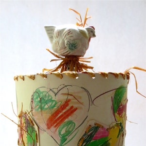 May include: A white ceramic chicken figurine sits atop a lampshade with a woven straw base. The lampshade is decorated with colorful crayon drawings of hearts.