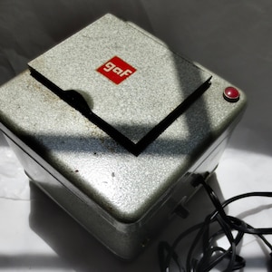 May include: A vintage silver-toned GAF slide projector with a red power button. The projector has a textured surface and a red square logo that says "gaf". A black power cord is connected to the projector.