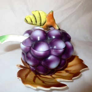 May include: A ceramic grape-shaped container with a lid, painted in shades of purple and white. It features a green and yellow leaf and a brown leaf-shaped base. The container is designed to resemble a bunch of grapes.