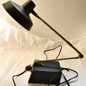 May include: A black desk lamp with a conical shade and an adjustable arm. The lamp has a rectangular base and a power cord. The arm is made of metal and has a brass-colored finish. The lamp is designed for desk use.