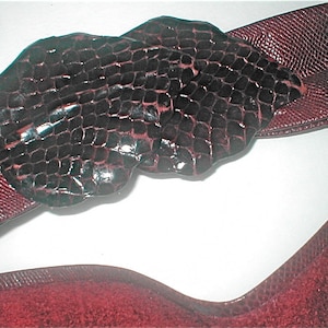 May include: A burgundy belt with a textured, dark brown buckle. The belt and buckle have a reptile-like pattern. The buckle is a unique shape, adding a distinctive touch to the accessory. The belt is a classic fashion item.