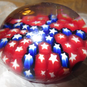 May include: A spherical glass paperweight featuring a dense millefiori pattern of red, white, and blue star shapes. The clear glass dome magnifies the internal arrangement of numerous small, colorful star motifs embedded throughout the solid object.
