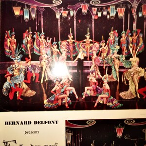 May include: A vintage theater program page featuring a large group of performers in colorful, elaborate stage costumes with ruffled skirts and headpieces, arranged on a dark stage under hanging lanterns, with the text Bernard Delfont presents.