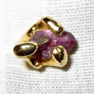 May include: A gold-colored jewelry piece with a rough, purplish-red gemstone at its center. The gold setting has a textured, organic design, with rounded, protruding elements. The gemstone appears to be a ruby.