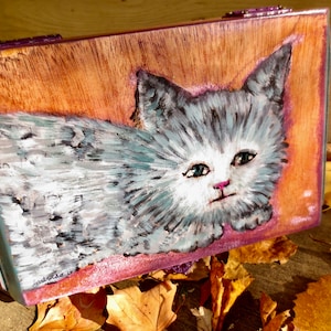 May include: Hand-painted wooden box featuring a gray and white kitten illustration. The kitten has blue eyes and a pink nose, set against a warm orange and purple background. The box is rectangular with a hinged lid and a purple trim.