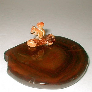 May include: A small, wooden, round trinket dish with a brown, orange, and white squirrel figurine sitting on a log on top.