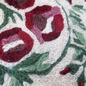 K&ouml;nnte beinhalten: Nahaufnahme eines cremefarbenen gestrickten Stoffes mit gestickten Blumen in Rot-, Rosa- und Gr&uuml;nt&ouml;nen.