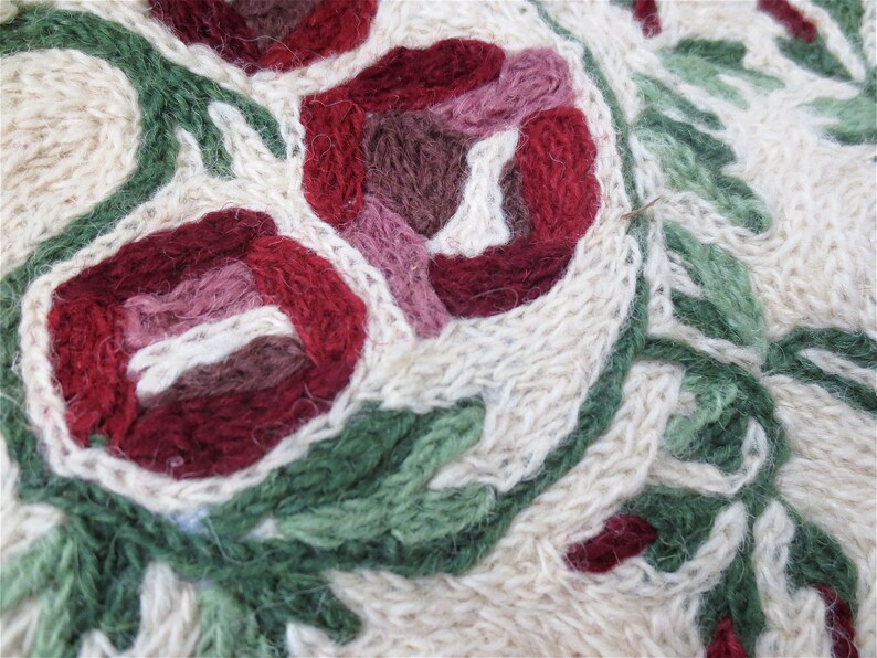 K&ouml;nnte beinhalten: Nahaufnahme eines cremefarbenen gestrickten Stoffes mit gestickten Blumen in Rot-, Rosa- und Gr&uuml;nt&ouml;nen.