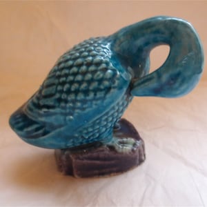 May include: A ceramic figurine of a duck in a teal glaze. The duck is perched on a dark purple base and is preening its feathers. The duck's body has a textured, scale-like pattern.