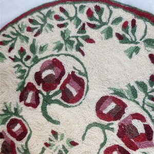 K&ouml;nnte beinhalten: Ein runder, cremefarbener Teppich mit einem floralen Muster. Der Teppich ist mit roten und gr&uuml;nen Blumen und Bl&auml;ttern bestickt.