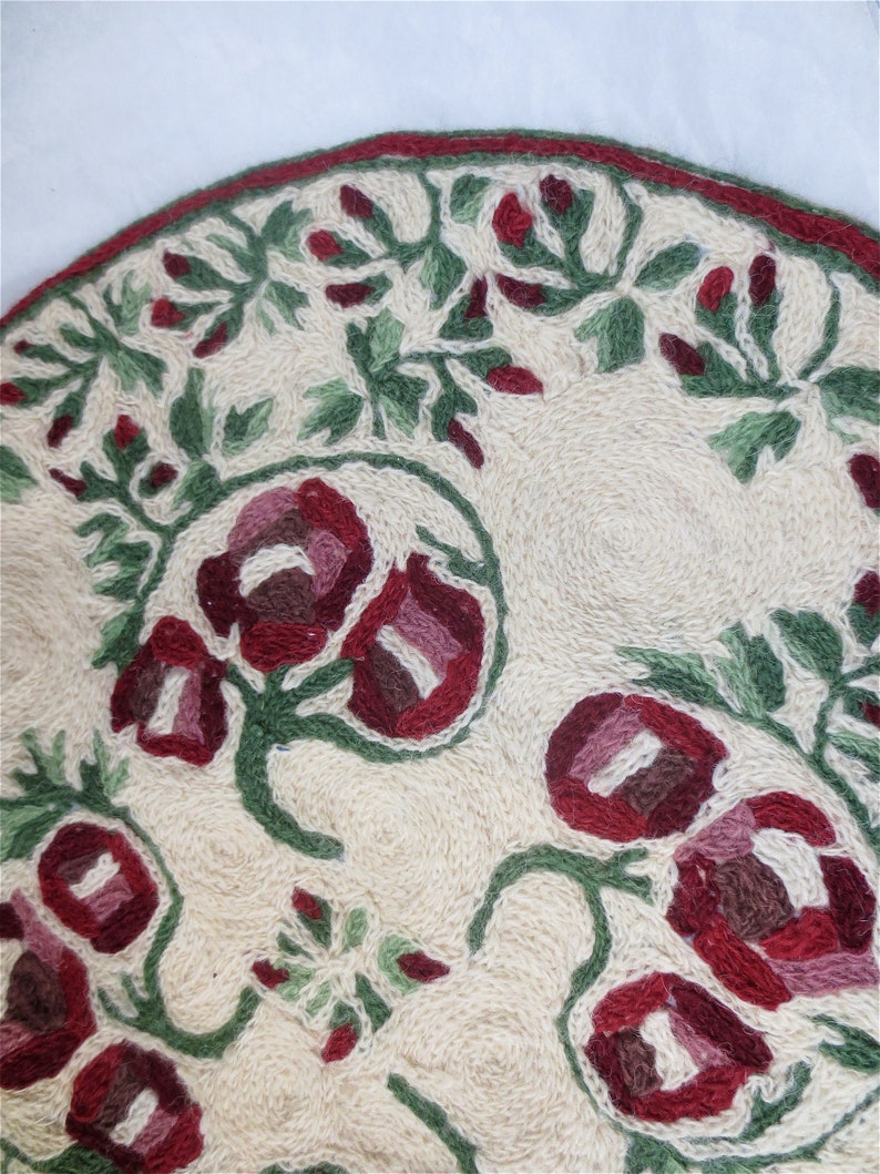 K&ouml;nnte beinhalten: Ein runder, cremefarbener Teppich mit einem floralen Muster. Der Teppich ist mit roten und gr&uuml;nen Blumen und Bl&auml;ttern bestickt.