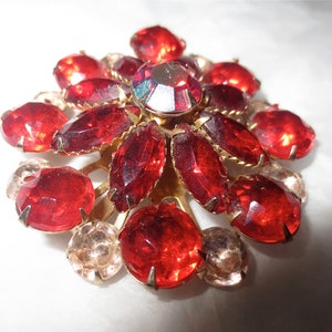 May include: A red and gold brooch with a large, central, faceted, clear rhinestone surrounded by smaller red rhinestones.