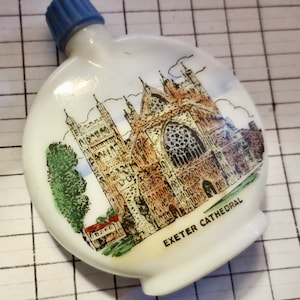 May include: White, oval-shaped flask with a blue cap. The flask features a detailed illustration of Exeter Cathedral, with the text "EXETER CATHEDRAL" below the image. The illustration includes the cathedral's architecture, trees, and sky.
