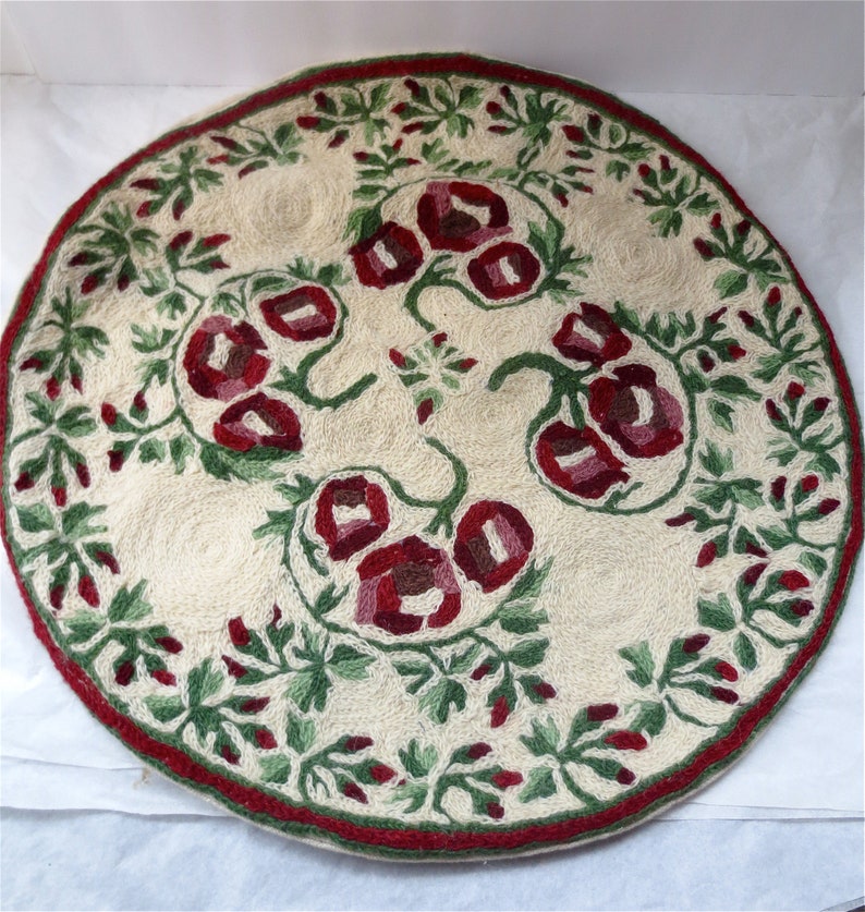 K&ouml;nnte beinhalten: Ein runder, cremefarbener Teppich mit einem roten und gr&uuml;nen Blumendesign. Der Teppich ist aus gewebtem Stoff und hat einen Fransenrand.