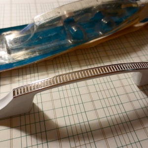 May include: White cabinet door handle with a chrome strip and a grooved design. The handle is in a plastic packaging.