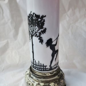 May include: A white glass vase with a black silhouette design of a tree, a fence, and a person playing a flute. The vase sits on a decorative, ornate metal base with a distressed, off-white finish.