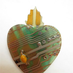 May include: Heart-shaped pendant with a circuit board design. The pendant features green and brown hues with copper-colored lines and a small, orange component. The top has a leaf-like accent with gold and clear sections.