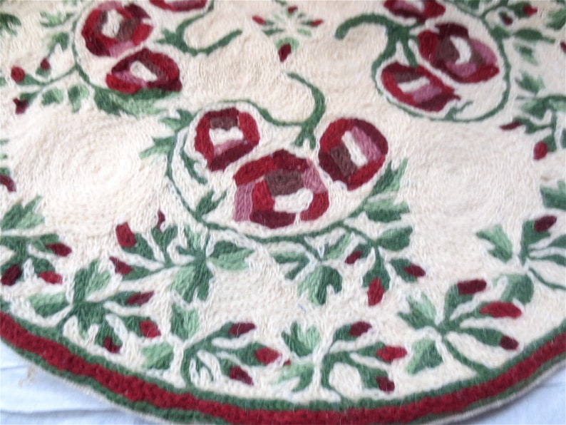 K&ouml;nnte beinhalten: Ein runder, cremefarbener Teppich mit einem roten und gr&uuml;nen Blumenmuster. Der Teppich hat einen roten Rand.