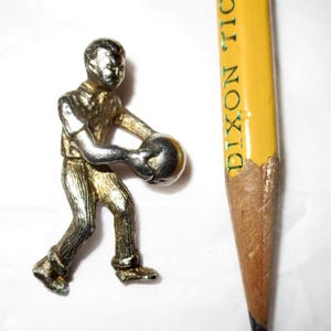 May include: A metallic silver-toned figurine of a person in a bowling stance holding a ball, positioned next to a yellow Dixon Ticonderoga pencil for scale. The figure features textured clothing details and a smooth, rounded bowling ball.