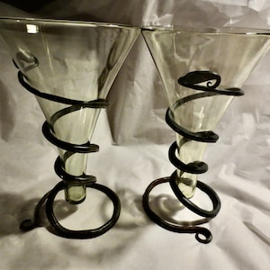May include: Two clear glass martini glasses with a dark metal spiral design wrapping around the stem and bowl. The base of each glass has a circular metal design. The glasses are on a white surface.