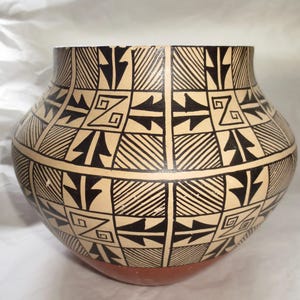 May include: A rounded ceramic pot featuring a cream-colored base with a repeating black geometric pattern. The design consists of alternating squares filled with diagonal lines and stylized arrow shapes, with a solid terracotta-colored base.