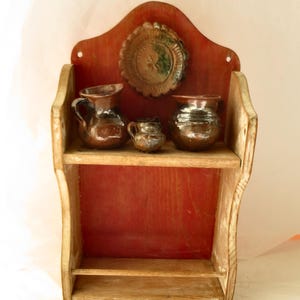 May include: A wooden wall shelf with a red back panel featuring a scalloped top edge. The shelf holds three small, dark brown glazed ceramic pitchers and a circular, scalloped-edge ceramic plate mounted on the back panel above the shelf.