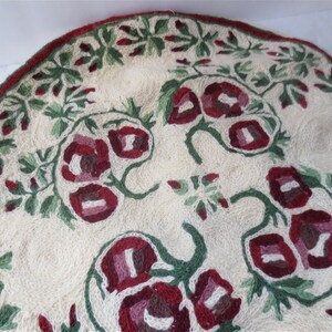 K&ouml;nnte beinhalten: Ein runder, cremefarbener Teppich mit einem floralen Muster. Der Teppich ist mit roten, rosa und gr&uuml;nen Blumen und Bl&auml;ttern bestickt.