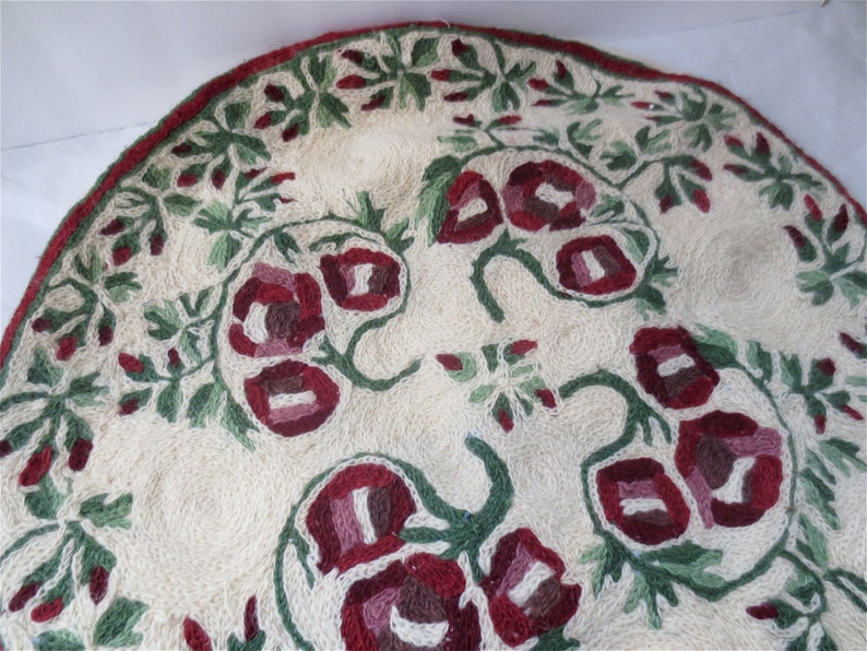 K&ouml;nnte beinhalten: Ein runder, cremefarbener Teppich mit einem floralen Muster. Der Teppich ist mit roten, rosa und gr&uuml;nen Blumen und Bl&auml;ttern bestickt.