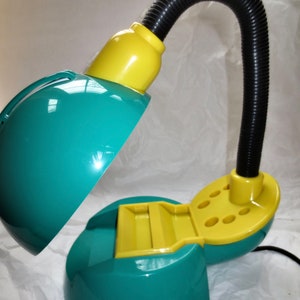 May include: A teal and yellow desk lamp with a flexible neck and a built-in storage compartment. The lamp has a dome-shaped shade and a round base.