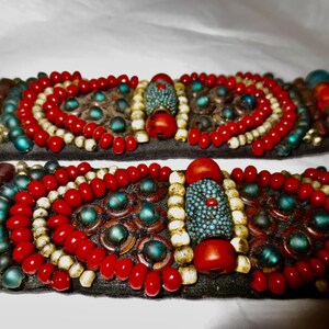 May include: Two parallel bands of black fabric adorned with intricate beadwork. The design features rows of vibrant red, cream, and teal beads, with a central textured teal cylindrical bead flanked by smooth red and cream spherical beads.