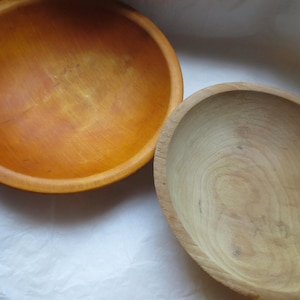 May include: Two round wooden bowls are shown from a top-down perspective. One bowl features a warm, golden-orange finish with a visible grain pattern, while the other bowl is a lighter, natural pale wood color with a distinct circular grain texture.