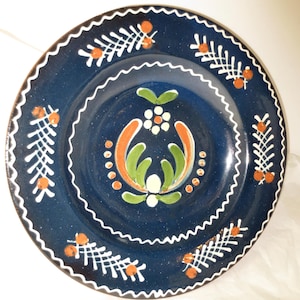 May include: A round, dark blue ceramic plate with white, orange, and green floral designs. The plate has a scalloped edge with white detailing and orange accents. The center features a stylized floral motif. A decorative piece for the home.