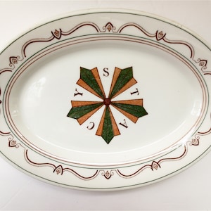 May include: Oval white ceramic serving platter with a decorative border and a central starburst design. The border features a brown and green wavy line with floral accents. The starburst has green and orange segments with the letters "Y", "C", "A", "T", and "S".