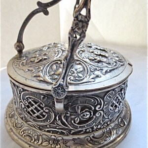 May include: A round silver-toned metal container with a hinged handle and a lid. The exterior features intricate embossed floral patterns, scrollwork, and lattice designs. The surface has a tarnished finish with dark recessed areas highlighting the detailed relief work on the lid and the sides of the base.