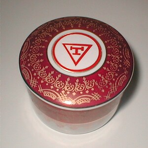 May include: A red and gold porcelain trinket box with a white circle containing a red triangle with a gold letter "T" inside. The box has a decorative pattern around the edge.